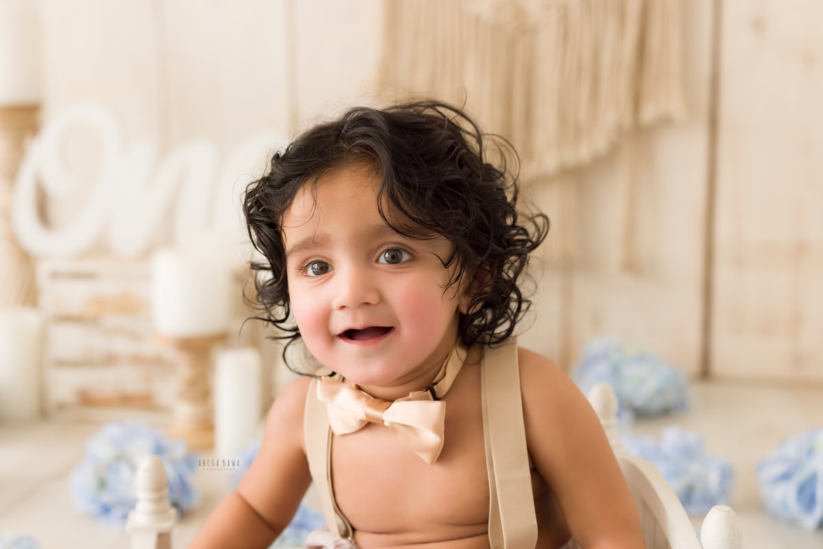 Embark on a visual journey through a baby's pre-birthday photoshoot in Delhi, featuring a dapper one-year-old boy adorned with a charming bow, surrounded by delicate blue flowers against a serene beige backdrop, meticulously captured by Anega Bawa in Gurgaon, Noida.