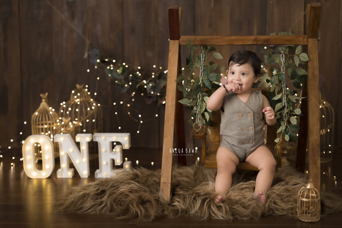Step into a fairy tale with your 1-year-old boy, swaying gently on a wooden swing amidst a cozy setting. With a brown rug underfoot, fairy lights twinkling, golden castles gleaming, and leafy fringes adorning the wall, let Anega Bawa Photography in Delhi and Gurgaon capture these magical moments through their expert sitter photography services.