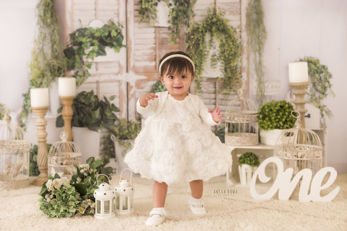 Capture the innocence and charm of a 1-year-old girl adorned in an all-white dress amidst a backdrop of candle stands and pots, set against a serene beige backdrop during a toddler photoshoot in Delhi, Gurgaon, and Noida by Anega Bawa.