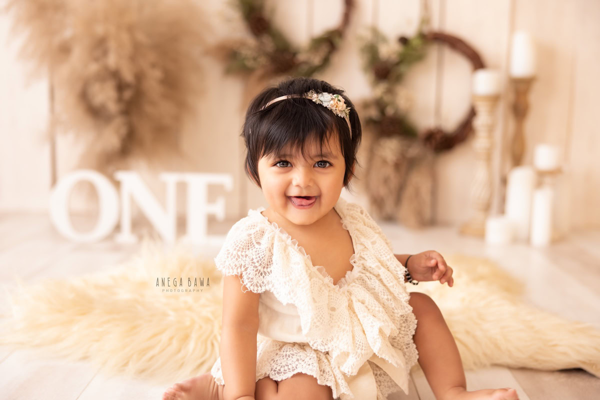 Capture the charm of a 1-year-old girl sitting on a beige rug, surrounded by candle stands, bushes, and a floral frame on a beige backdrop. Perfect for a sitter photography session in Delhi and Gurgaon by Anega Bawa.
