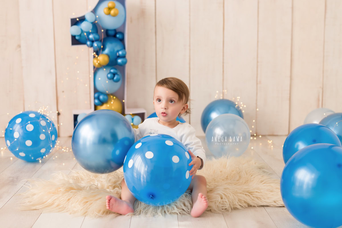 Immerse in the delightful sight of a 1-year-old girl surrounded by blue balloons against a soothing beige backdrop, complemented by a cozy beige rug during a toddler photoshoot in Delhi, Gurgaon, and Noida by Anega Bawa.