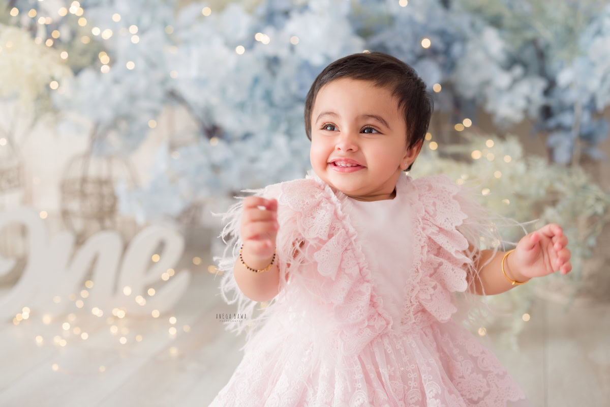 Captivating one-year-old girl surrounded by delicate blue flowers and twinkling fairy lights against a floral backdrop, capturing the magic of her first birthday photoshoot in Delhi, by Anega Bawa, Gurgaon, Noida