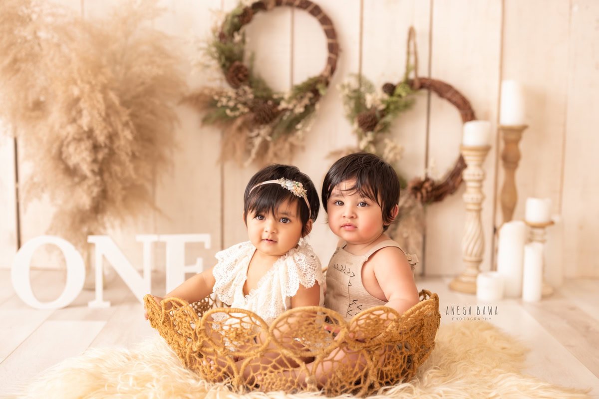 Adorable 1-year-old girl and boy in a rustic brown basket, surrounded by elegant candle stands, floral wooden frames on the wall, lush brown bushes, set against a warm beige backdrop, captured by professional sitter photographer Anega Bawa in Gurgaon, Delhi.