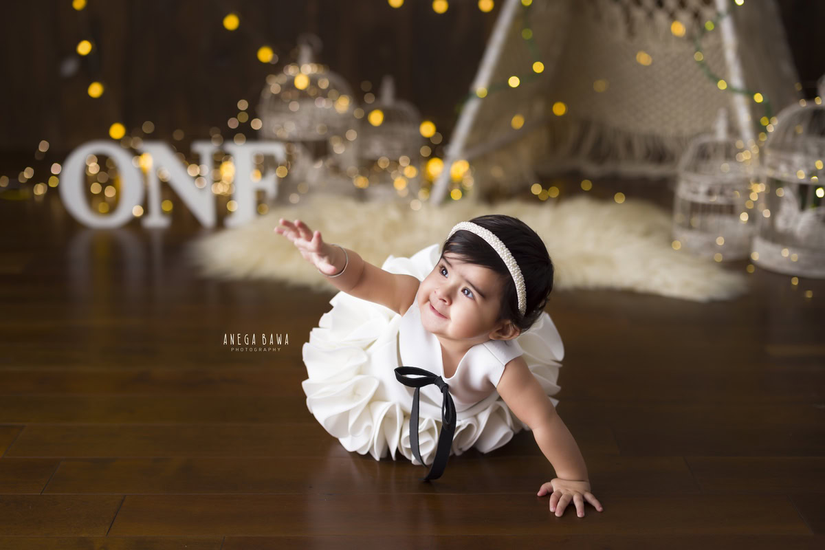 Witness the charm of a crawling 1-year-old girl, exploring the world within a net tent, adorned with a white castle and twinkling fairy lights, all set against a warm beige rug. This captivating moment captured during a toddler photoshoot in Delhi, Gurgaon, and Noida by Anega Bawa is pure magic.