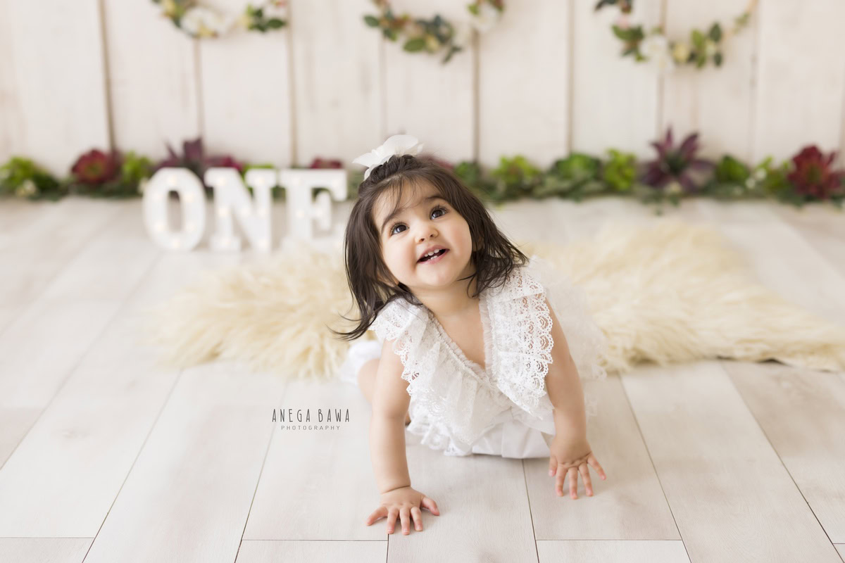 Experience the delightful sight of a 1-year-old girl joyfully exploring her surroundings, crawling on a cozy beige rug. The scene is adorned with wooden floral frames adorning the walls, creating a picturesque setting. This charming moment was captured during a toddler photoshoot in Delhi, Gurgaon, and Noida by Anega Bawa.