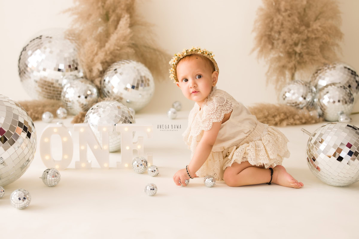 Celebrate the joy of movement with a 1-year-old girl as she explores her surroundings, crawling amidst discoballs and bushes against a serene beige backdrop. Ideal for a delightful sitter photoshoot in Delhi and Gurgaon by Anega Bawa.