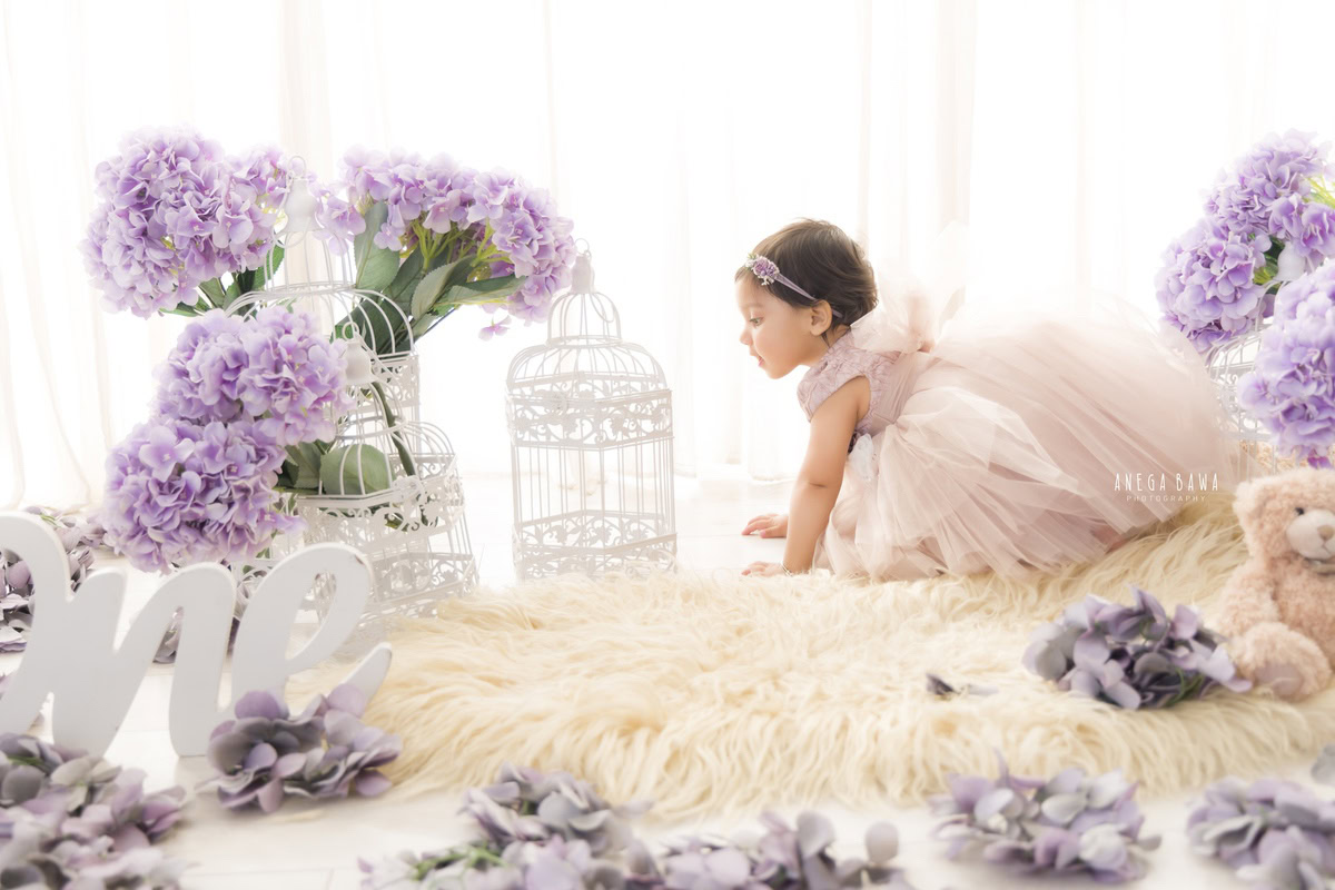 Witness the adorable sight of a 1-year-old girl gleefully exploring her surroundings, crawling on a soft beige rug. Surrounding her are whimsical white castles and delicate lavender flowers against a pristine white backdrop. This precious moment was immortalized during a toddler photoshoot in Delhi, Gurgaon, and Noida by the talented Anega Bawa.