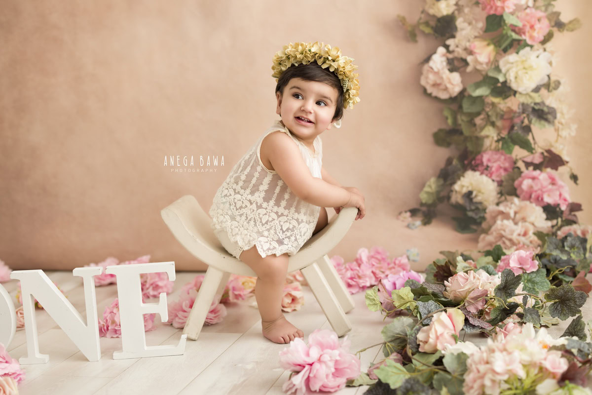 Capture the charm of a 1-year-old girl seated on a curvy white chair against a soft peach backdrop adorned with a delicate floral fringe and a dainty tiara band. Perfect for a memorable sitter photoshoot in Delhi and Gurgaon by Anega Bawa.