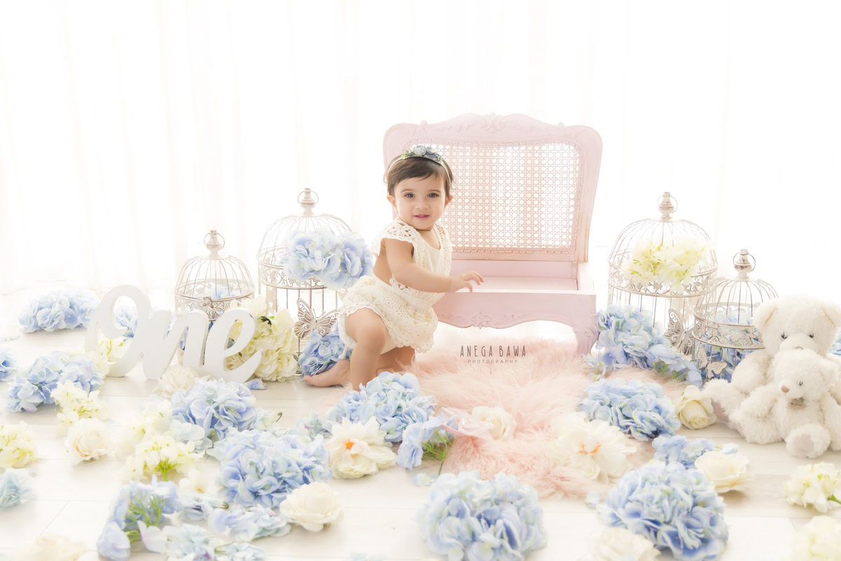 Watch your 1-year-old princess hold court in a whimsical photoshoot by Anega Bawa Photography in Delhi and Gurgaon. Clutching a pink wooden chair amidst a backdrop of white castles and a pink rug strewn with blue and white flowers, she creates enchanting memories in every frame.