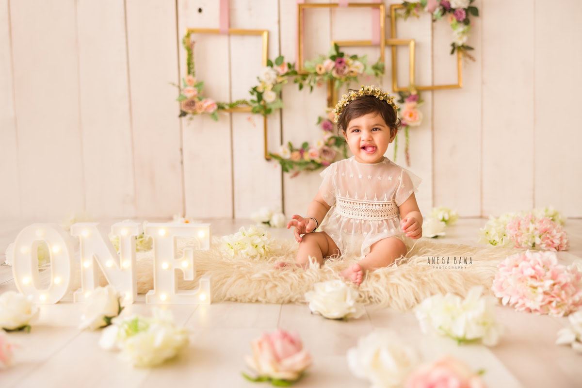 Envision a precious moment with a 1-year-old girl in a charming net dress, seated on a cozy beige rug amidst scattered flowers. Behind her, square floral frames adorn the wall against a serene beige backdrop, creating an enchanting scene. Perfect for capturing the innocence and beauty of childhood in a sitter photoshoot in Delhi and Gurgaon by Anega Bawa.