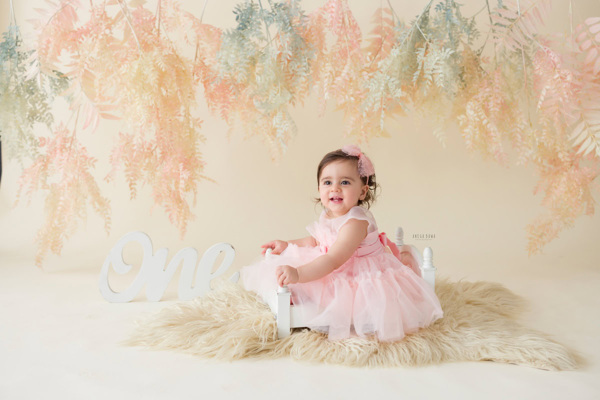 Delight in capturing your one-year-old princess in a charming photoshoot with Anega Bawa Photography in Delhi and Gurgaon. Picture her nestled in a white cot atop a beige rug against a backdrop adorned with leafy accents in soothing peach hues, while wearing an adorable headband that adds to her angelic charm.