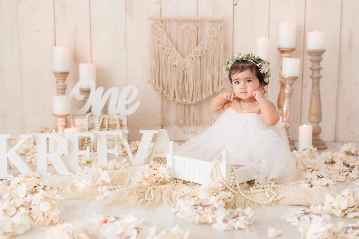Experience the magic of your one-year-old princess captured in a delightful sitter photoshoot by Anega Bawa Photography in Delhi and Gurgaon. Watch her shine in a white net dress, surrounded by the warm glow of candlelight and framed by her name on the floor against a serene beige backdrop.