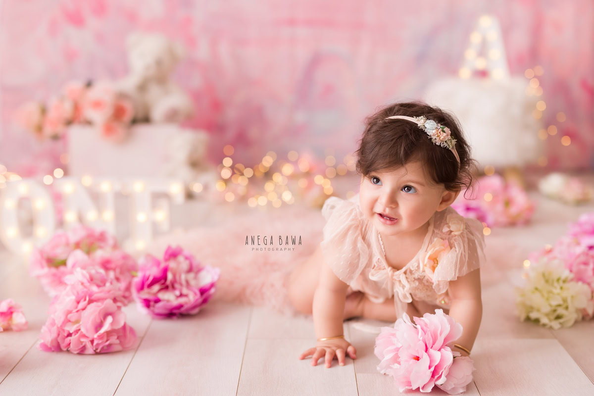 Capture the enchantment of your 1-year-old girl in a sitter photoshoot by Anega Bawa Photography in Delhi and Gurgaon. Your little one sits on a beige floor adorned with pink flowers, against a pink backdrop illuminated by fairy lights, creating a magical and memorable scene.
