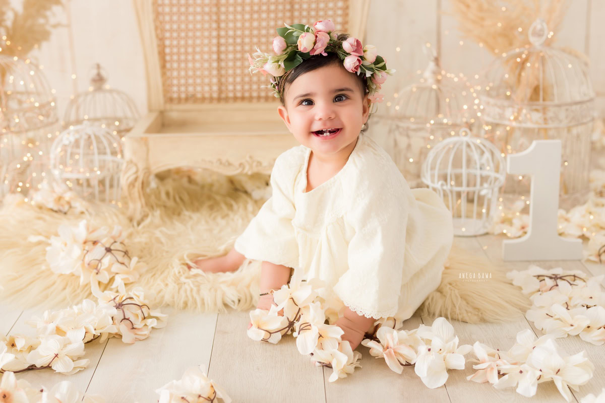 Step into the enchanting realm of a baby's pre-birthday photoshoot in Delhi, where a darling one-year-old girl graces a beige rug adorned with a sparkling tiara band, set against a backdrop of pristine white castles against a serene beige backdrop, expertly captured by Anega Bawa in Gurgaon, Noida.
