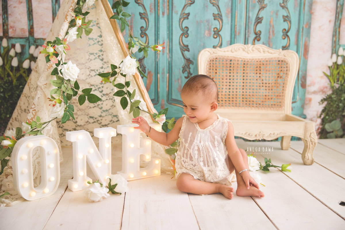 Charming 1-year-old girl playing with a decorative frame on the floor, surrounded by leafy fringes, near a cozy tent, set against a serene blue backdrop, captured by professional sitter photographer Anega Bawa in Gurgaon, Delhi.
