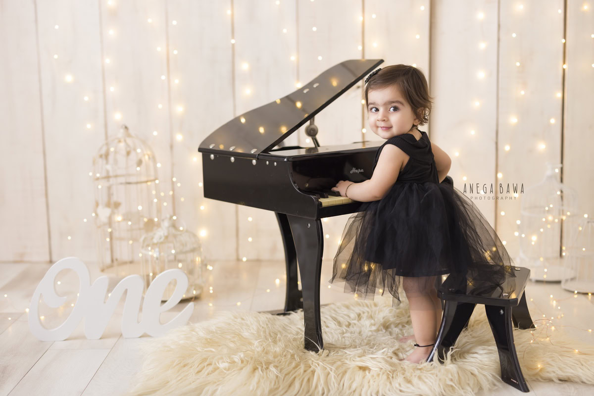 Adorable 1-year-old girl playing a piano on a beige rug, illuminated by fairy lights, with a dreamy beige backdrop and castles, captured by talented sitter photographer Anega Bawa in Gurgaon, Delhi.