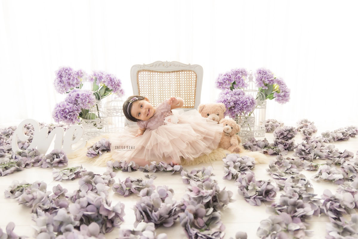 Witness the adorable charm of your one-year-old princess in a captivating sitter photoshoot by Anega Bawa Photography in Delhi and Gurgaon. See her striking a pose of pure sweetness on a cute wooden chair, with delicate flowers adorning the floor, all set against a pristine white backdrop.