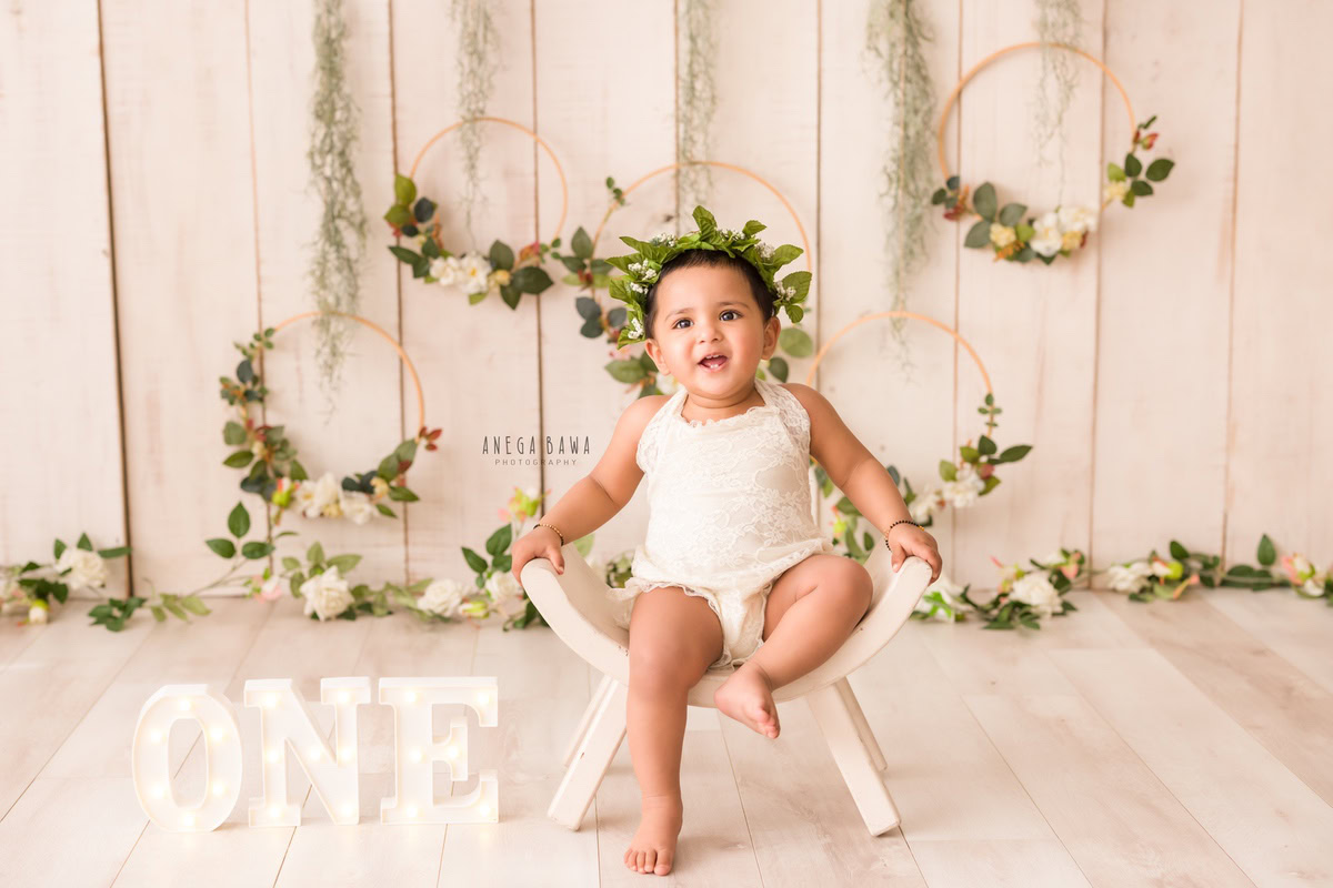 Immerse yourself in the sweetness of a 1-year-old girl seated on a curvy chair, set against a soothing beige backdrop. Adorning the wall are leafy wooden frames, adding a touch of natural elegance to the scene. It's an ideal moment for a sitter photoshoot in Delhi and Gurgaon, skillfully captured by Anega Bawa.