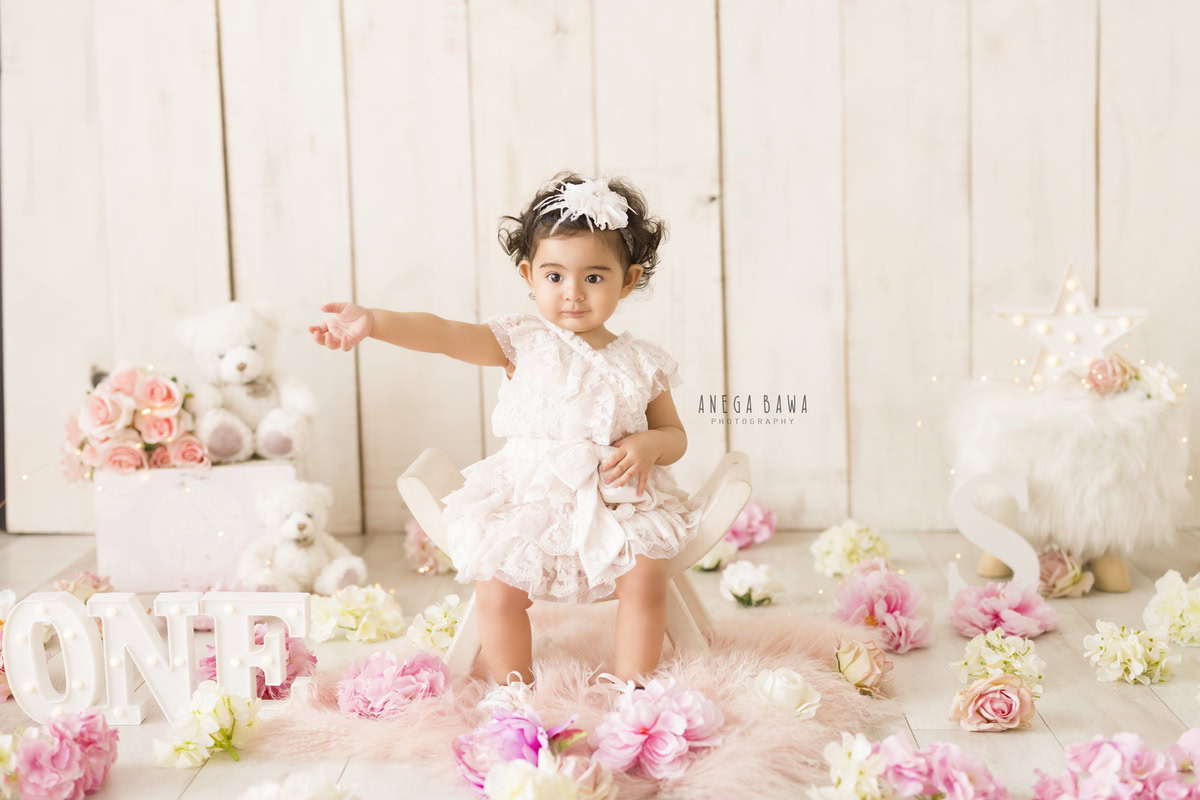 Charming 1-year-old girl seated on a curvy chair with a peach rug, against a beige backdrop adorned with multicolored flowers and a cute headband, captured by expert sitter photographer Anega Bawa in Gurgaon, Delhi.