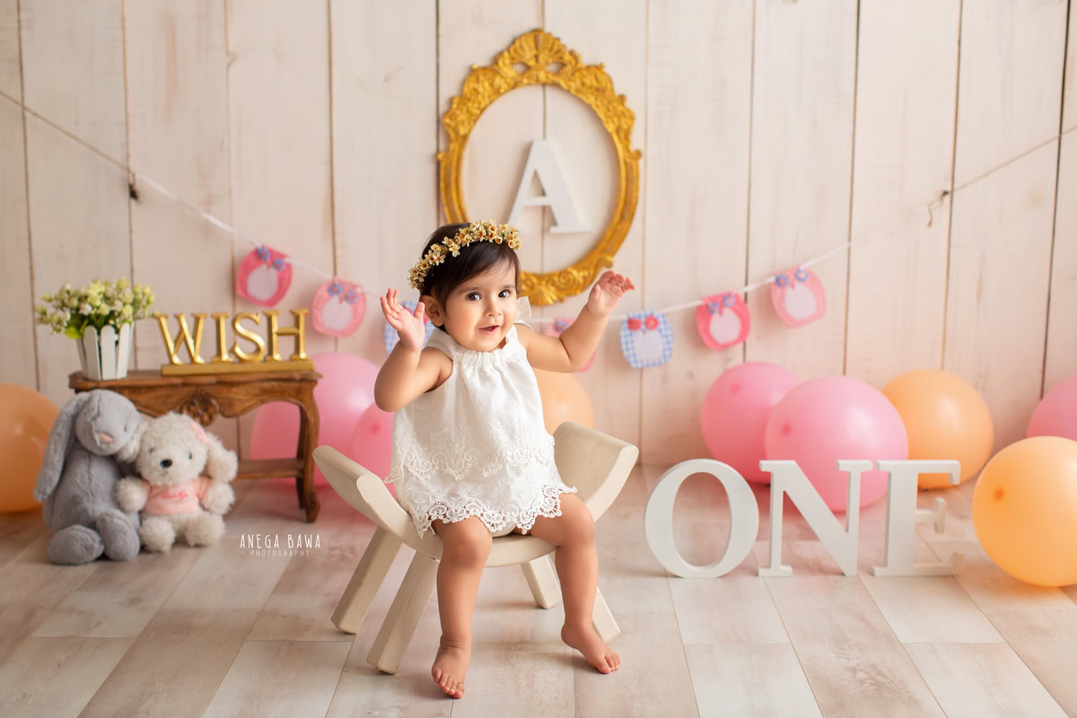 Capture the charm of your 1-year-old girl in a sitter photoshoot by Anega Bawa Photography in Delhi and Gurgaon. She sits on a curvy chair surrounded by teddybears and balloons, with an alphabet frame on the wall, all set against a beige backdrop, creating an adorable and memorable scene.