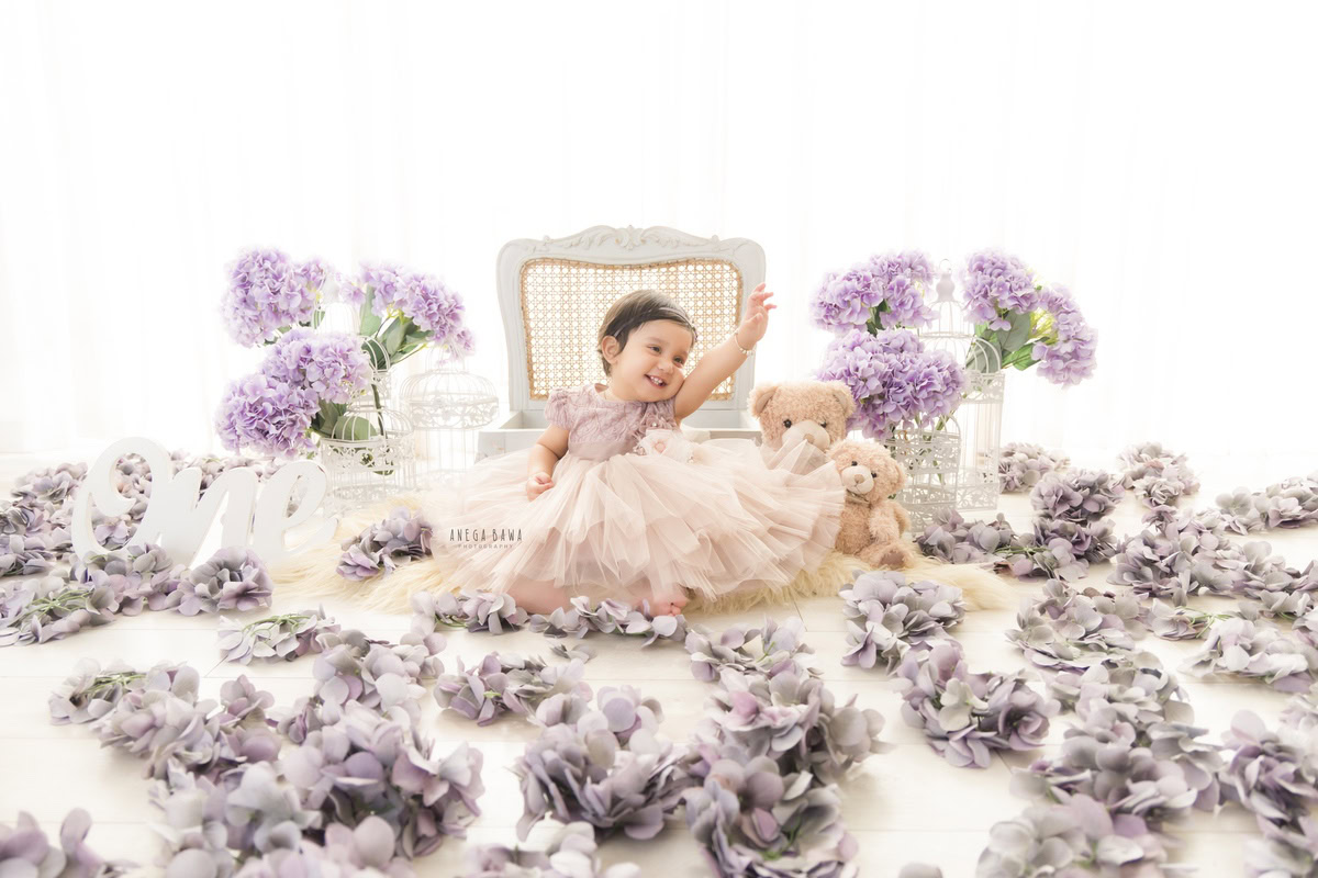 Capture the delightful innocence of your one-year-old princess in a captivating sitter photoshoot by Anega Bawa Photography in Delhi and Gurgaon. See her seated gracefully on the floor amidst scattered flowers, her presence illuminated against a serene white backdrop. A wooden chair and a charming flower vase add to the enchanting ambiance, creating timeless memories to cherish.