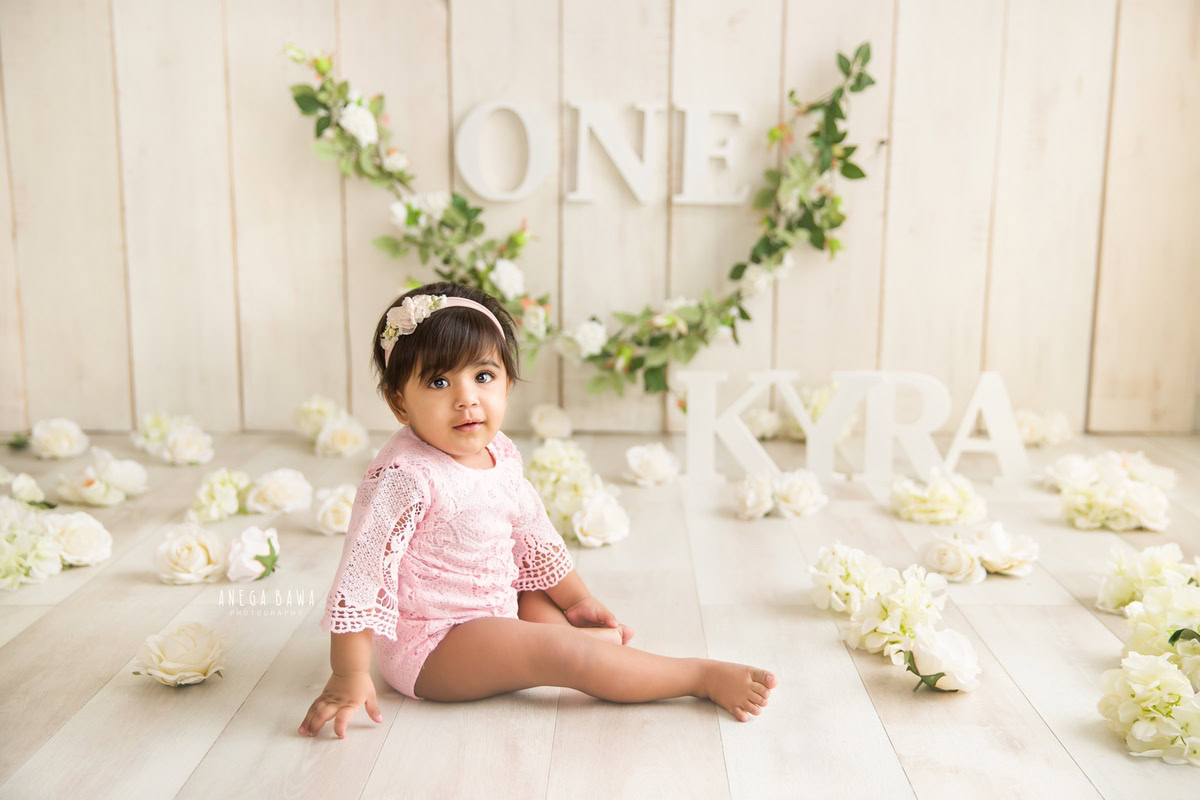Witness the serenity of a 1-year-old girl seated amidst delicate white flowers, against a warm beige backdrop. It's a captivating scene captured for a sitter photoshoot in Delhi and Gurgaon, expertly orchestrated by Anega Bawa.