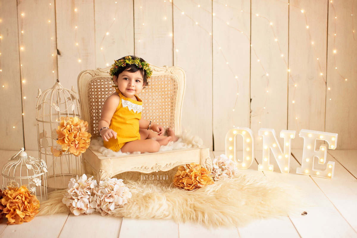 Capture the precious moments of your 1-year-old girl with Anega Bawa Photography in Delhi and Gurgaon. She sits gracefully on a wooden chair with a tiara band, surrounded by orange and white flowers and fairy lights, all set against a beige backdrop with a beige rug, creating a beautiful and timeless sitter photoshoot.