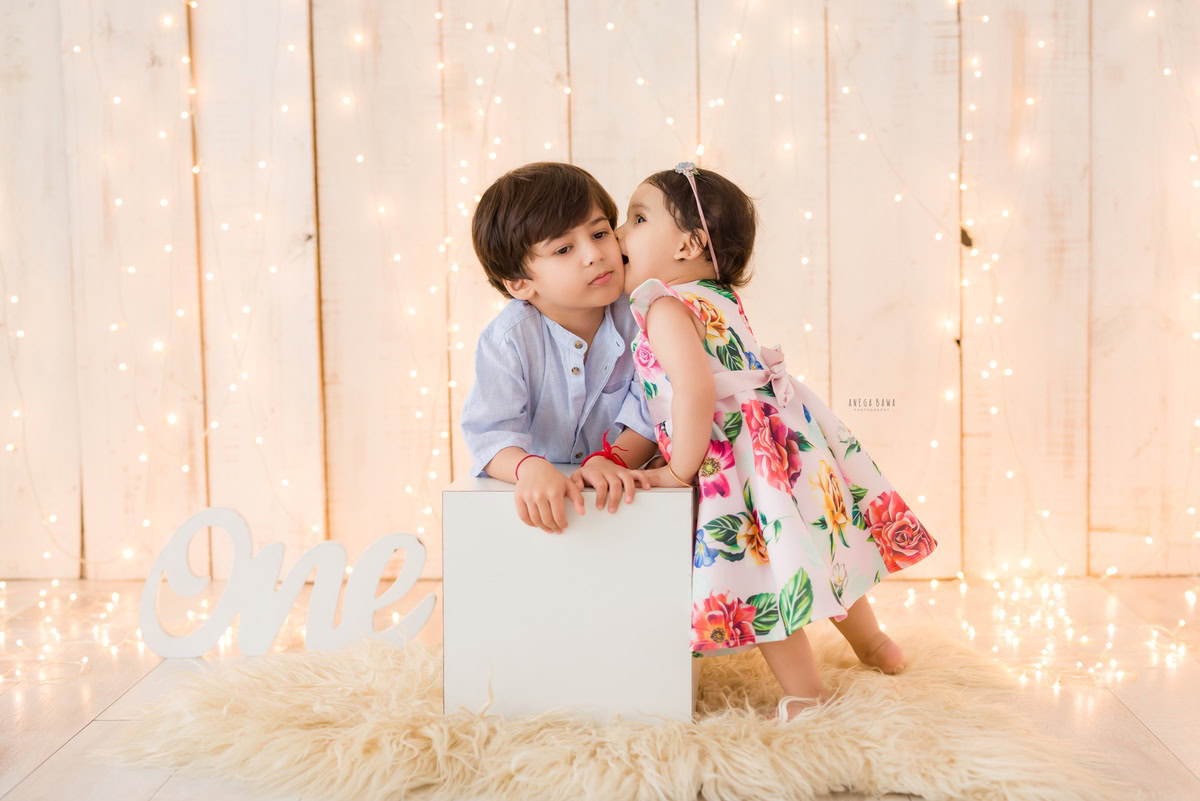 Experience the heartwarming charm of a baby's pre-birthday photoshoot in Delhi, featuring an adorable one-year-old girl sharing a tender sibling kiss on a cozy beige rug against a soft beige backdrop, skillfully captured by Anega Bawa in Gurgaon, Noida.