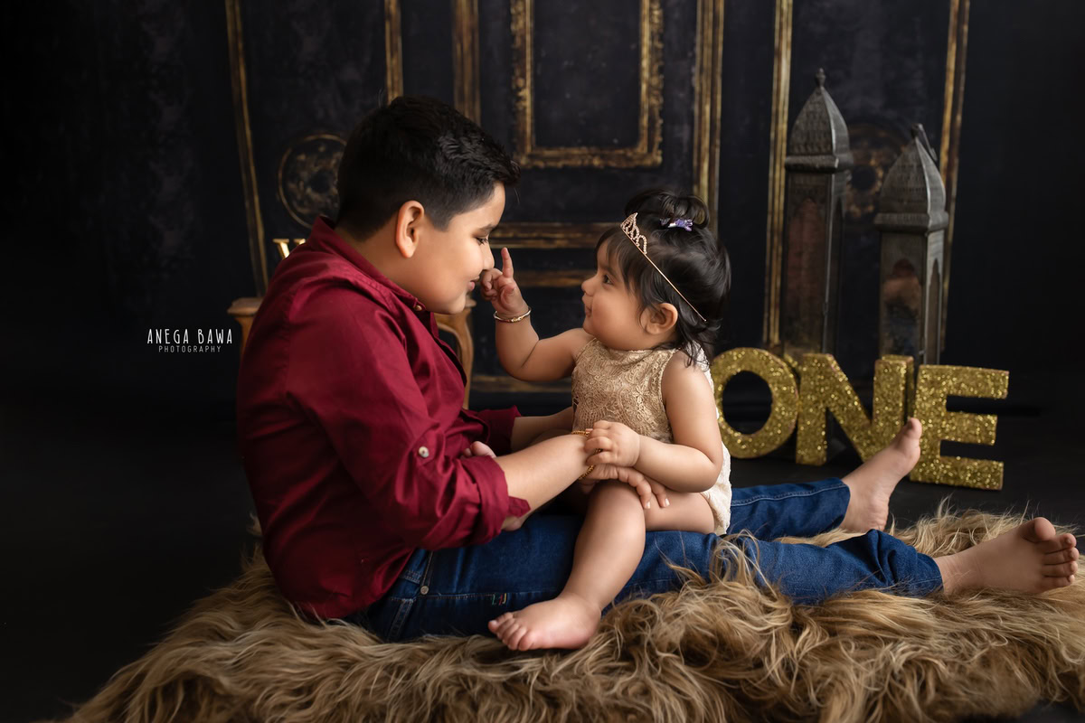 Capture the precious moments of your 1-year-old girl and her sibling with Anega Bawa Photography in Delhi and Gurgaon. They sit together on a brown rug against a brown backdrop, creating a warm and timeless setting for a beautiful sitter photoshoot.