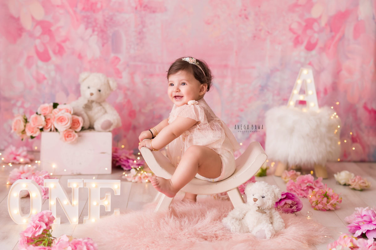 Capture the innocence and charm of a 1-year-old girl seated on a curvy chair, surrounded by a delightful pink backdrop adorned with flowers. A plush pink rug and a cuddly teddy bear complete the enchanting scene. This heartwarming moment is expertly preserved through a sitter photoshoot in Delhi and Gurgaon, courtesy of Anega Bawa.