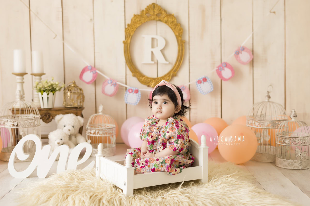 Delight in the innocence captured as a 1-year-old girl sits snugly in a white cot, surrounded by enchanting elements. Adorning the wall is an alphabet frame, adding a touch of education to the scene. The soft beige rug beneath her feet complements the setting, along with whimsical castles and a delicate candle stand. This charming moment was expertly preserved during a toddler photoshoot in Delhi, Gurgaon, and Noida by the talented Anega Bawa.