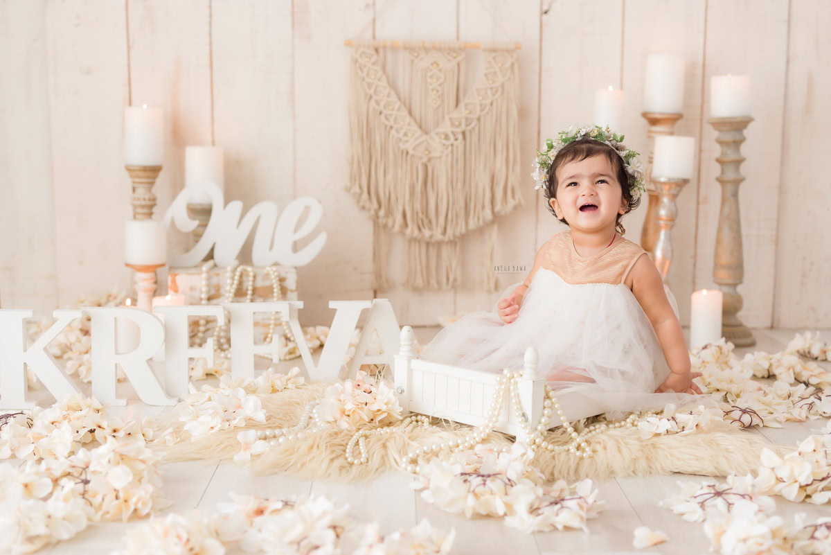 Celebrate your one-year-old princess with a charming sitter photoshoot by Anega Bawa Photography in Delhi and Gurgaon. Adorned in a dainty tiara band, she sits gracefully in a white cot, surrounded by candlelight against a pristine white backdrop. The beige rug adds warmth, while delicate flowers and a name frame on the floor enhance the enchanting ambiance. Let Anega Bawa Photography capture these precious moments for timeless memories.