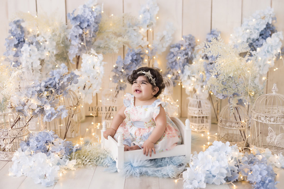 Celebrate your little princess's journey to one year with a captivating sitter photoshoot in Delhi and Gurgaon by Anega Bawa Photography. Nestled in a cozy white cot, surrounded by the soft glow of fairy lights against a serene beige backdrop, she's adorned with delicate blue and white flowers, creating enchanting memories. Trust Anega Bawa Photography for your one-year-old photoshoot in Delhi and Gurgaon, capturing precious moments you'll treasure forever.