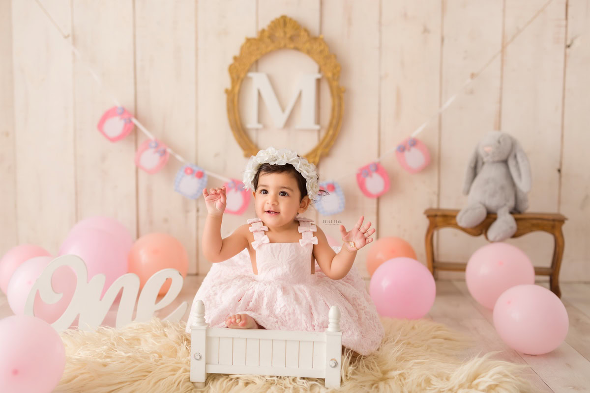 Sweet one-year-old girl sitting in a pristine white cot adorned with pink balloons and teddy bears, framed by alphabet letters on the wall, set upon a cozy beige rug against a serene beige backdrop, capturing the charm of her first birthday photoshoot in Delhi, by Anega Bawa, Gurgaon, Noida