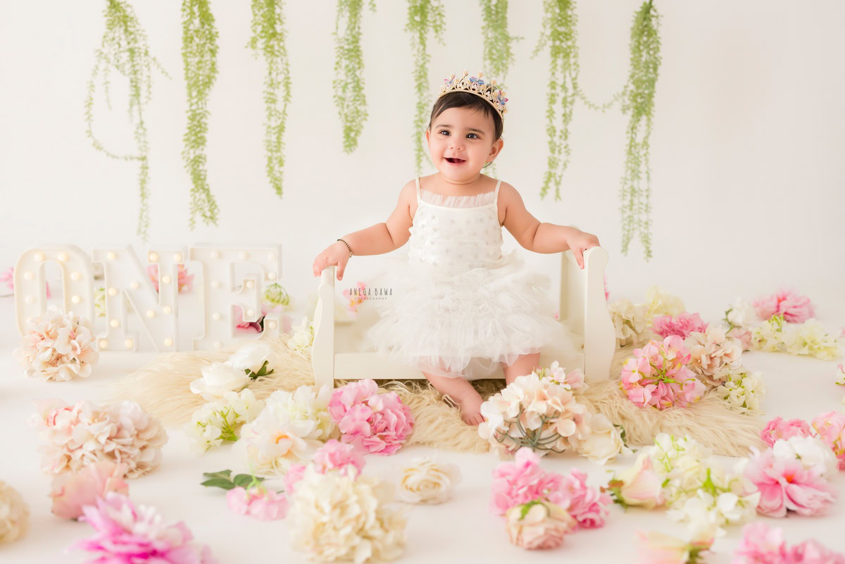 Immerse yourself in the delightful world of a baby's pre-birthday photoshoot in Delhi, where a precious one-year-old girl sits gracefully in a white cot amidst a bed of pink and white flowers, crowned with a leafy fringe crown headband, expertly captured by Anega Bawa in Gurgaon, Noida.