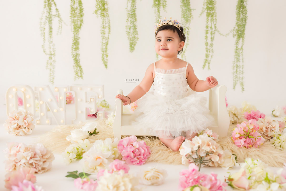 Capture the pure joy of your little one with a charming sitter photography session in Delhi and Gurgaon by Anega Bawa Photography. Adorably seated in a pristine white cot, surrounded by a delicate arrangement of pink and white flowers and leafy fringes, against a serene white backdrop, your one-year-old creates timeless memories. Trust Anega Bawa Photography for a delightful sitter photoshoot experience in Delhi and Gurgaon, preserving precious moments to cherish for a lifetime.