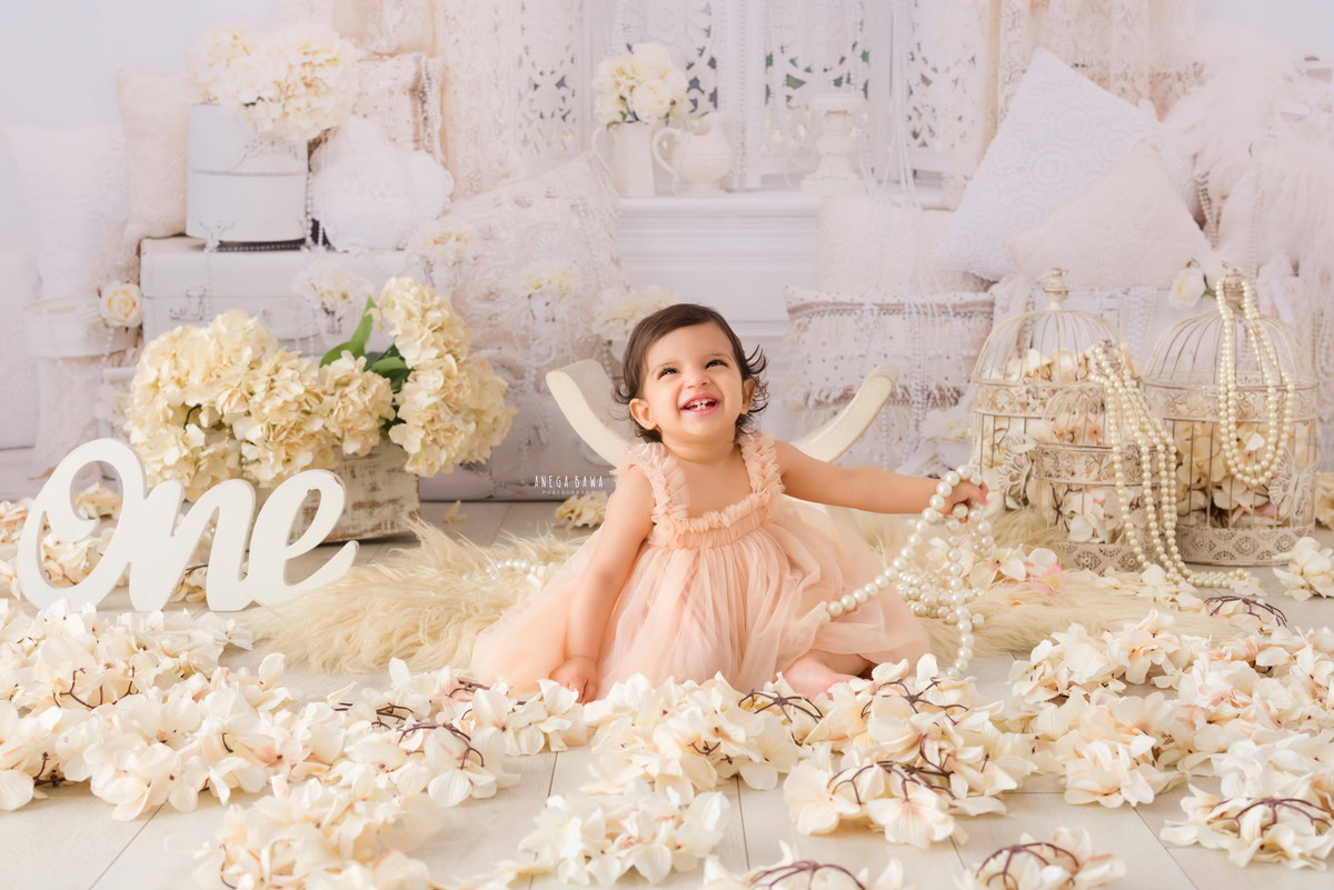 Adorable one-year-old girl sitting on a cozy beige rug surrounded by enchanting castles and delicate pearls amidst white flowers, against a pristine white backdrop, commemorating her magical first birthday photoshoot in Delhi, by Anega Bawa, Gurgaon, Noida