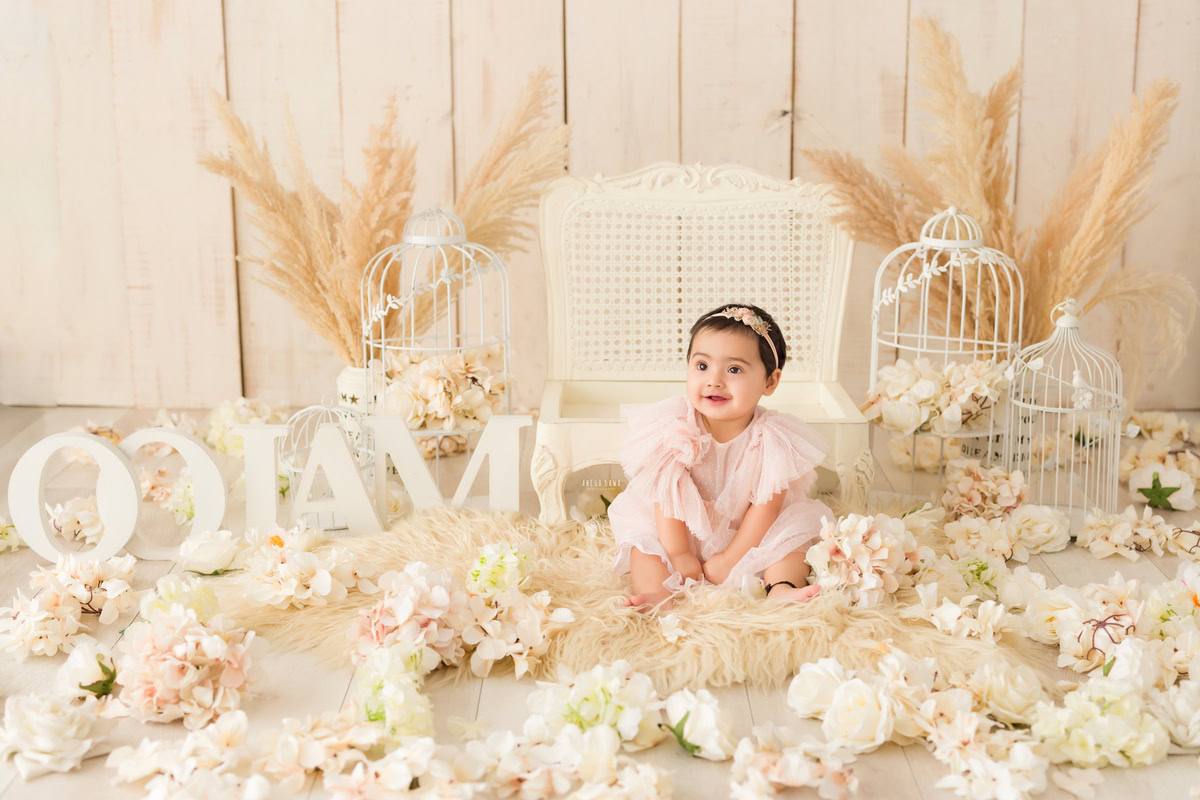 Sweet one-year-old girl sitting on a cozy beige rug amidst a charming name frame on the floor, embellished with delicate white flowers, bushes, and castles, against a serene beige backdrop, capturing the enchantment of her first birthday photoshoot in Delhi, by Anega Bawa, Gurgaon, Noida