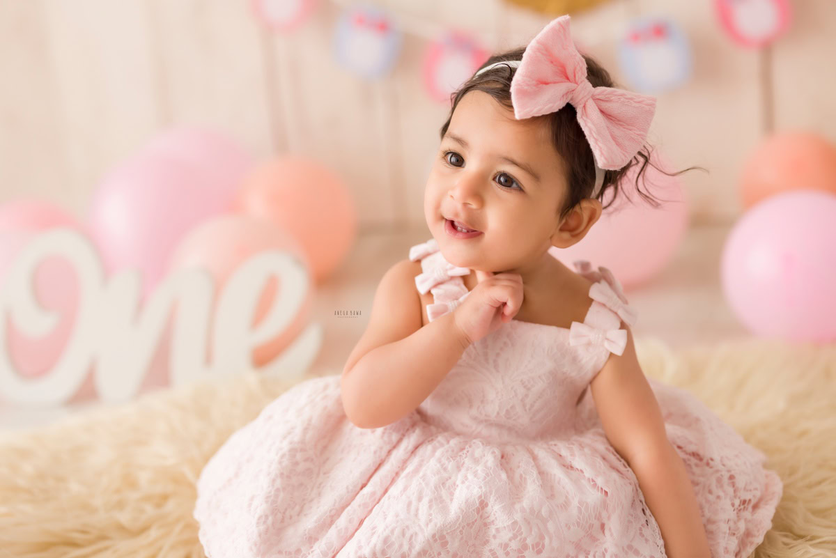 Immerse yourself in the innocence and joy as a 1-year-old girl sits gracefully on a beige rug, surrounded by a whimsical display of peach and pink balloons against a serene beige backdrop. Adorned with a cute headband, she radiates pure happiness in this delightful moment captured during a toddler photoshoot in Delhi, Gurgaon, and Noida by the talented Anega Bawa.