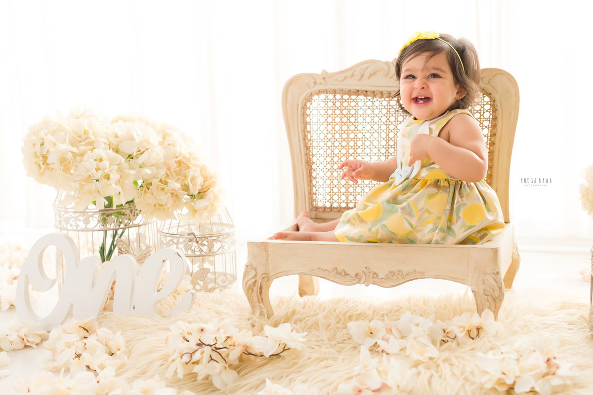 Witness the charm of a 1-year-old girl seated gracefully on a chair, surrounded by delicate ivory flowers and a pristine white rug. Against a backdrop of pure white, with whimsical castles adding a touch of magic, this enchanting scene was captured during a tender toddler photoshoot in Delhi, Gurgaon, and Noida by the talented Anega Bawa.