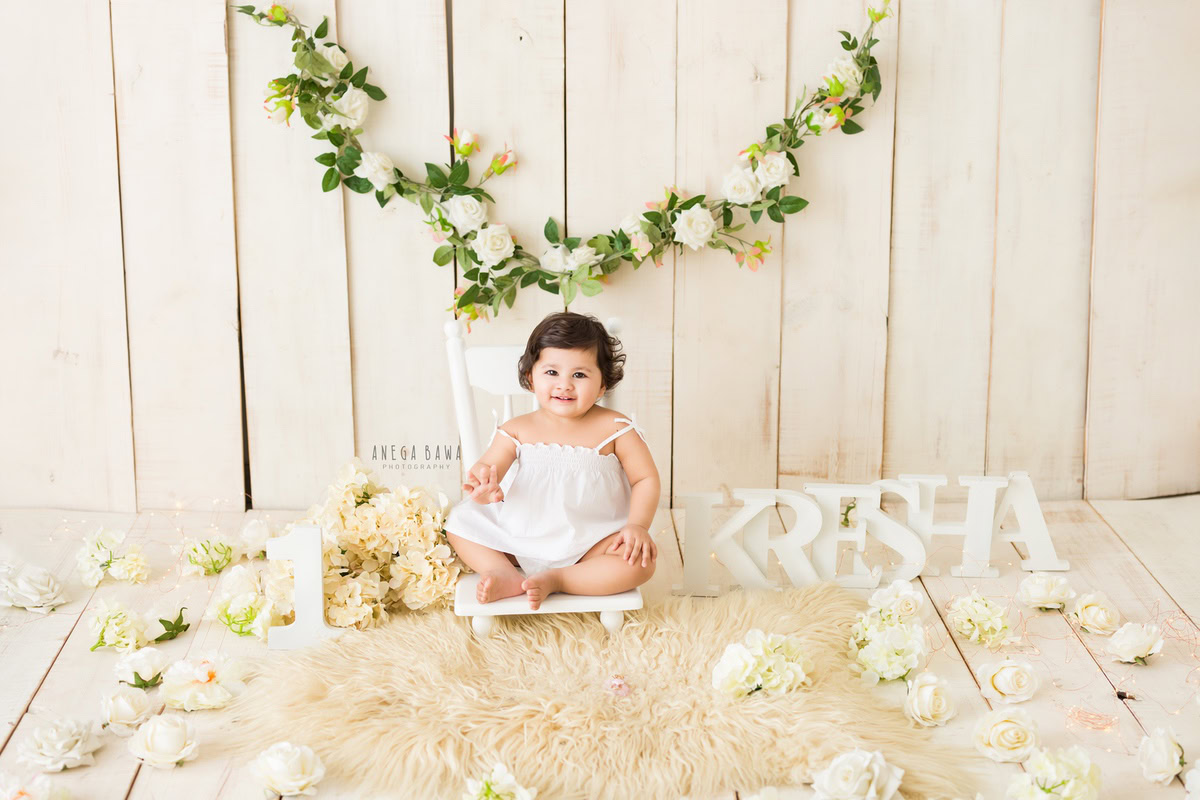 Create unforgettable memories of your 1-year-old girl with Anega Bawa Photography in Delhi and Gurgaon. Watch her charm shine as she sits on a white chair atop a beige rug, surrounded by flowers on the floor, against a beige backdrop adorned with a leafy garland on the wall, making for a delightful sitter photoshoot.