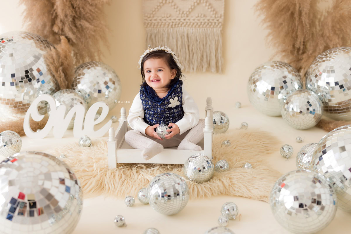 Embark on a journey through the whimsical wonderland of a baby's pre-birthday photoshoot in Delhi, as a charming one-year-old girl sits atop a white cot, surrounded by twinkling disco balls and lush bushes against a soothing beige backdrop, skillfully captured by Anega Bawa in Gurgaon, Noida.