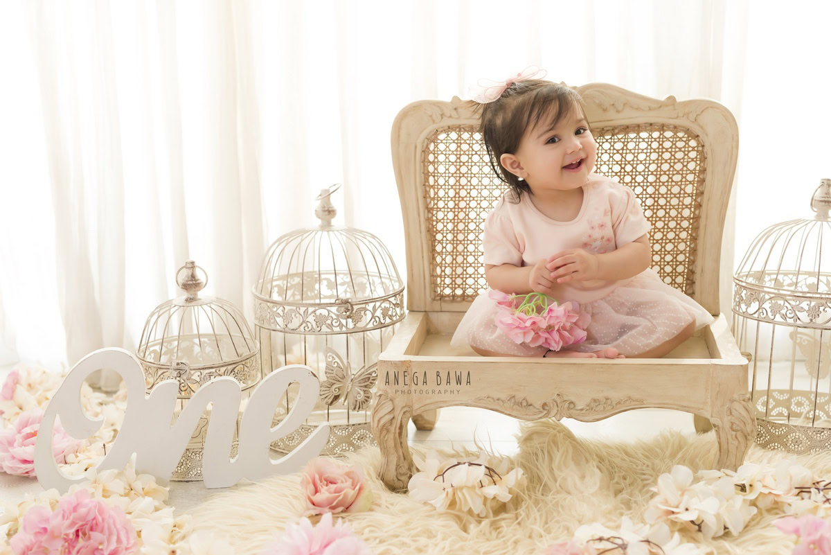 Experience the adorable innocence of a 1-year-old girl seated on a wooden chair, surrounded by white castles and delicate pink and white roses. Against a backdrop of pure white, this charming moment was beautifully captured during a toddler photoshoot in Delhi, Gurgaon, and Noida by the skilled photographer, Anega Bawa.