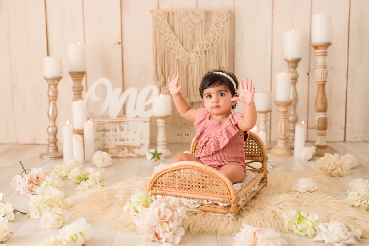 Step into the enchanting realm of a baby's pre-birthday photoshoot in Delhi, where a delightful one-year-old girl perches on a wooden cot, accompanied by elegant candle stands and delicate white flowers, set against a backdrop of beige rug and beige backdrop, skillfully crafted by Anega Bawa in Gurgaon, Noida.