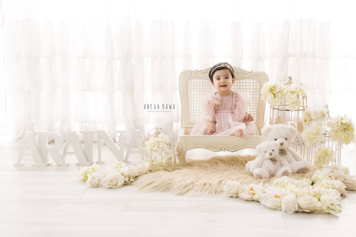 Adorable 1-year-old girl sitting against a white backdrop with a beige rug, surrounded by teddy bears and castles, captured by talented sitter photographer Anega Bawa in Gurgaon, Delhi.