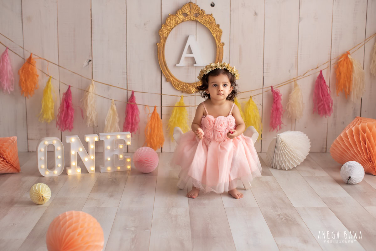 Charming 1-year-old girl standing in front of a golden frame with alphabet letters, wall hangings, and colorful pompom decorations, set against a beige backdrop, beautifully photographed by Anega Bawa, sitter photographer in Gurgaon, Delhi.
