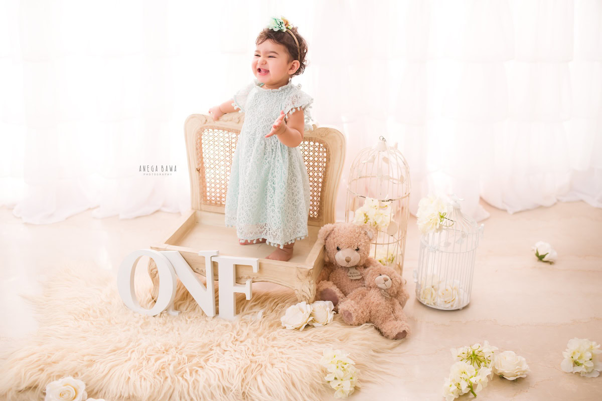 Capture the innocence and joy of your 1-year-old girl with Anega Bawa Photography in Delhi and Gurgaon. Let her stand on a wooden chair placed on a beige rug, surrounded by white flowers and teddy bears, against a serene white backdrop for a memorable sitter photoshoot.