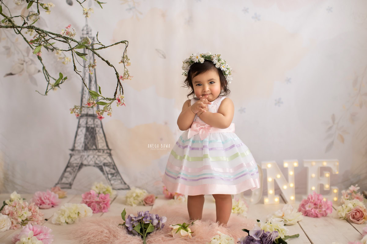 Immerse in the charm of your 1-year-old girl's sitter photoshoot with Anega Bawa Photography in Delhi and Gurgaon. Watch her stand gracefully on a pink rug against a backdrop adorned with an Eiffel Tower wallpaper, capturing moments filled with sweetness and wonder.