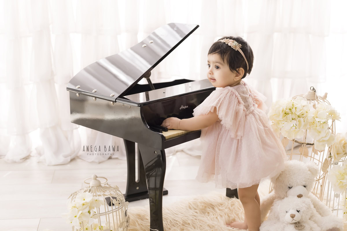 Adorable 1-year-old girl standing with a piano, castles, and teddybears on the floor, set against a pristine white backdrop, beautifully captured by Anega Bawa, sitter photographer in Gurgaon, Delhi.