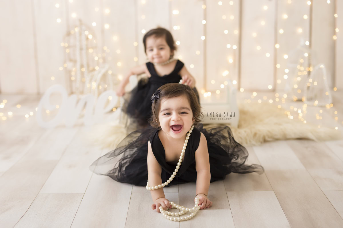 Adorable one-year-old twin girls in elegant black dresses adorned with pearls, against a soft beige backdrop, illuminated by twinkling fairy lights, capturing the magic of their first birthday photoshoot in Delhi, by Anega Bawa, Gurgaon, Noida